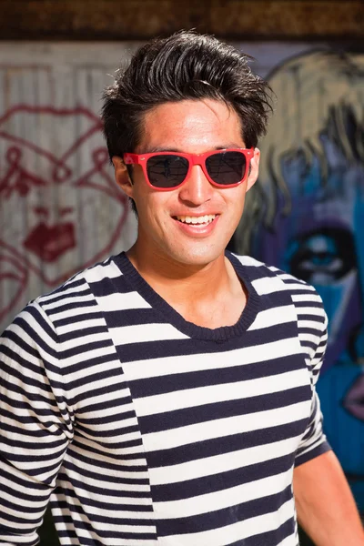 Urban asian man with red sunglasses. Good looking. Cool guy. Wearing blue white striped sweater. Standing in front of wooden wall with graffiti. — Stock Photo, Image