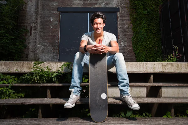 Ein urbaner asiatischer Mann mit Skateboard sitzt auf einer Treppe. gut aussehend. Cooler Typ. trägt graues Hemd und Jeans. altes, vernachlässigtes Gebäude im Hintergrund. — Stockfoto