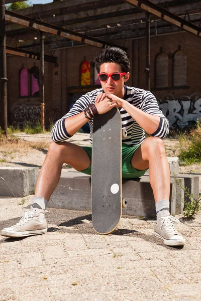 Urbaner asiatischer Mann mit roter Sonnenbrille und Skateboard auf der Straße. gut aussehend. Cooler Typ. Er trug einen blau-weiß gestreiften Pullover und eine grüne Shorts. altes, vernachlässigtes Gebäude im Hintergrund. — Stockfoto