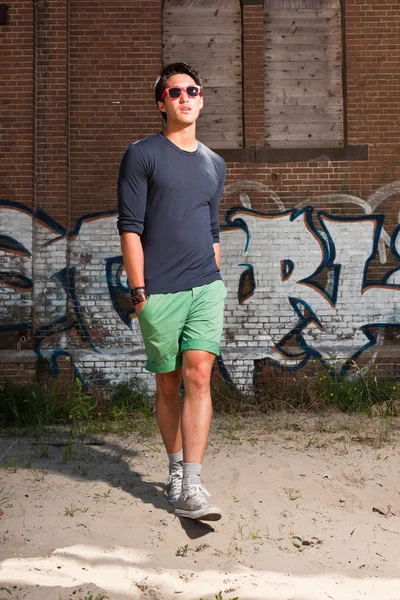 Urbano asiático homem com óculos de sol vermelhos. Boa aparência. Um tipo fixe. Vestindo camisa azul escuro e shorts verdes. Em pé na frente da parede de tijolo com graffiti . — Fotografia de Stock