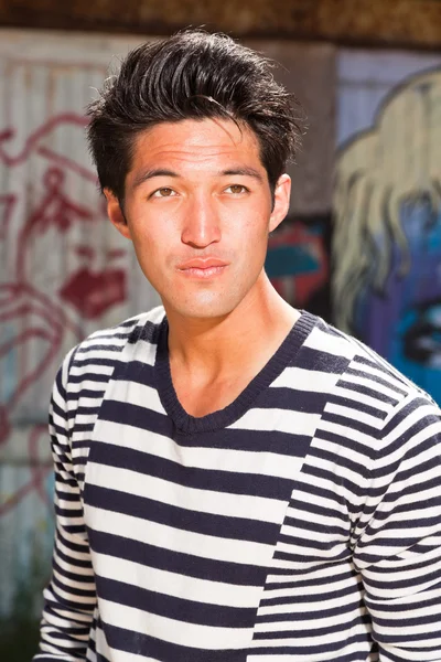 Homme asiatique urbain. Beau look. Cool mec. Portant un pull rayé bleu blanc. Debout devant un mur en bois avec des graffitis . — Photo