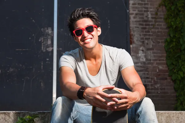 Urban asian man with red sunglasses and skateboard sitting on stairs. Good looking. Cool guy. Wearing grey shirt and jeans. Old neglected building in the background. — Stock Photo, Image