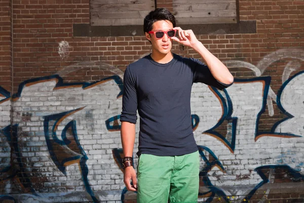 Urban uomo asiatico con occhiali da sole rossi. Bell'aspetto. Che figo. Indossa camicia blu scuro e pantaloncini verdi. In piedi davanti al muro di mattoni con graffiti . — Foto Stock