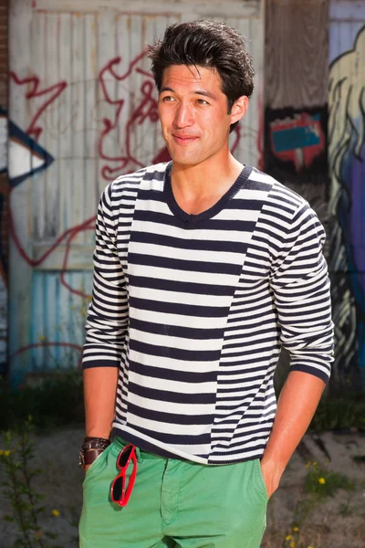 Hombre asiático urbano. Muy guapo. Un tipo genial. Usando jersey azul a rayas blancas y pantalones cortos verdes. De pie frente a la pared de madera con graffiti . —  Fotos de Stock