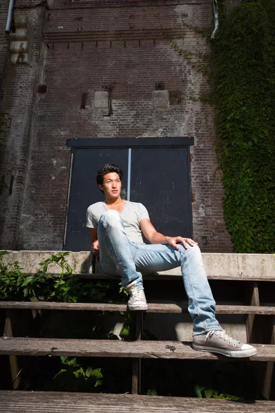 Urban asian man sitting on stairs. Good looking. Cool guy. Wearing grey shirt and jeans. Old neglected building in the background. — Stock Photo, Image