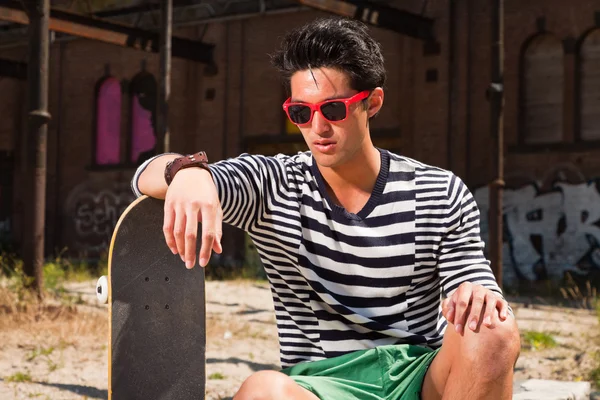 Urban asiatiska man med röda solglasögon och skateboard sitter på gatan. Snygg. cool kille. bär blå vit randig tröja och gröna shorts. gammal försummade byggnad i bakgrunden. — Stockfoto