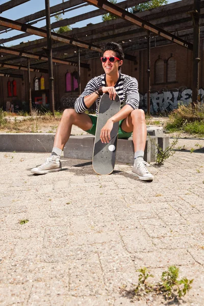Urban asian man with red sunglasses and skateboard sitting on street. Good looking. Cool guy. Wearing blue white striped sweater and green shorts. Old neglected building in the background. — Stock Photo, Image