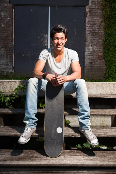 Urban asian man with skateboard sitting on stairs. Good looking. Cool guy. Wearing grey shirt and jeans. Old neglected building in the background. — Stock Photo, Image