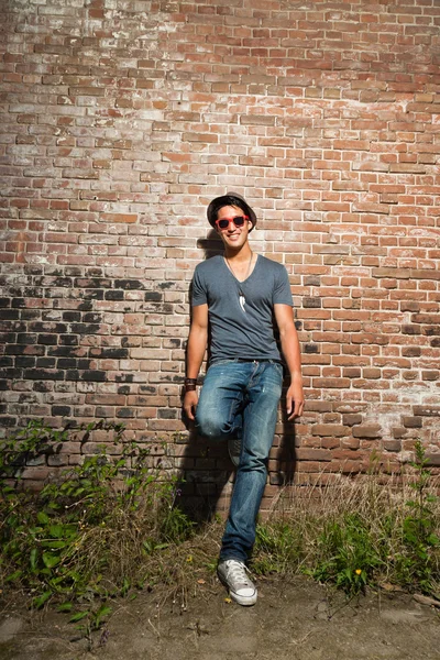 Urbano asiático homem com óculos de sol vermelhos. Boa aparência. Um tipo fixe. Vestindo camisa cinza e chapéu e jeans. Em pé na frente da parede de tijolo . — Fotografia de Stock