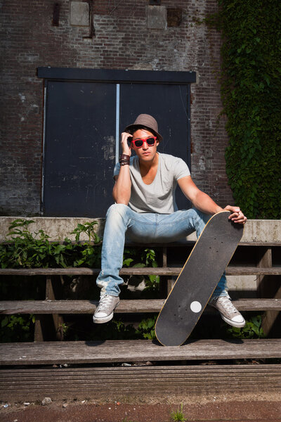 Urban asian man with hat, red sunglasses and skateboard sitting on stairs. Good looking. Cool guy. Wearing grey shirt and jeans. Old neglected building in the background.