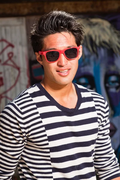 Hombre asiático urbano con gafas de sol rojas. Muy guapo. Un tipo genial. Usando jersey azul a rayas blancas. De pie frente a la pared de madera con graffiti . —  Fotos de Stock