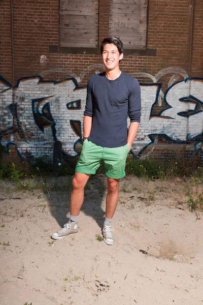 Urbano asiático homem. Boa aparência. Um tipo fixe. Vestindo camisa azul escuro e shorts verdes. Em pé na frente da parede de tijolo com graffiti . — Fotografia de Stock