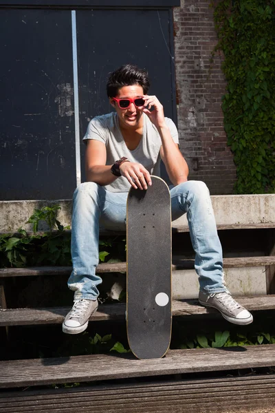 Ein urbaner asiatischer Mann mit roter Sonnenbrille und Skateboard sitzt auf einer Treppe. gut aussehend. Cooler Typ. trägt graues Hemd und Jeans. altes, vernachlässigtes Gebäude im Hintergrund. — Stockfoto