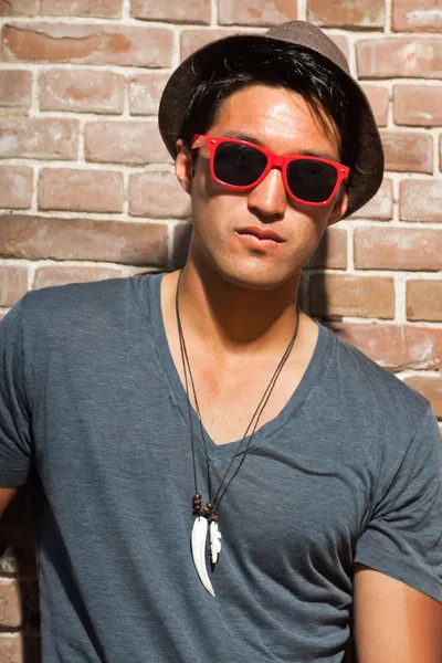Αστικές ασιατικό άτομο με κόκκινα γυαλιά ηλίου. Ευπαρουσίαστο. ωραίος τύπος. φορώντας γκρι πουκάμισο και καπέλο. στέκεται μπροστά από τον τοίχο από τούβλα. — Φωτογραφία Αρχείου