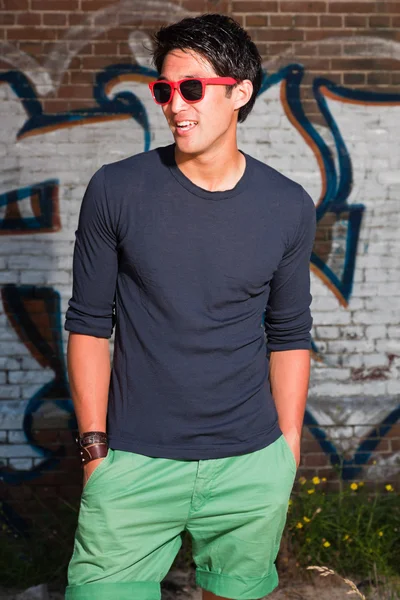 Hombre asiático urbano con gafas de sol rojas. Muy guapo. Un tipo genial. Con camisa azul oscuro y pantalones cortos verdes. De pie frente a la pared de ladrillo con graffiti . —  Fotos de Stock