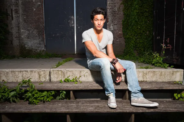 Urbaner asiatischer Mann auf einer Treppe sitzend. gut aussehend. Cooler Typ. trägt graues Hemd und Jeans. altes, vernachlässigtes Gebäude im Hintergrund. — Stockfoto