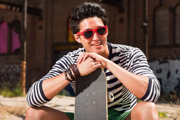 Urban asian man with red sunglasses and skateboard sitting on street. Good looking. Cool guy. Wearing blue white striped sweater and green shorts. Old neglected building in the background. — Stock Photo, Image