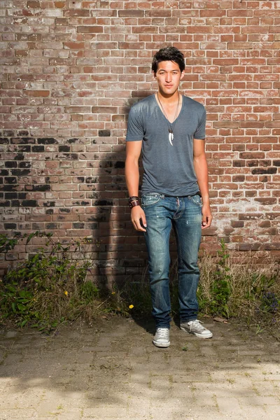 Hombre asiático urbano. Muy guapo. Un tipo genial. Usando camisa gris y jeans. De pie frente a la pared de ladrillo . — Foto de Stock