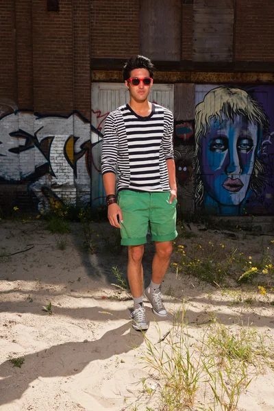 Hombre asiático urbano con gafas de sol rojas. Muy guapo. Un tipo genial. Usando jersey azul a rayas blancas y pantalones cortos verdes. De pie frente a la pared con graffiti . — Foto de Stock