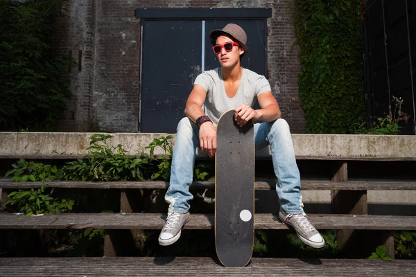 Urban asian man with hat, red sunglasses and skateboard sitting on stairs. Good looking. Cool guy. Wearing grey shirt and jeans. Old neglected building in the background. — Stock Photo, Image