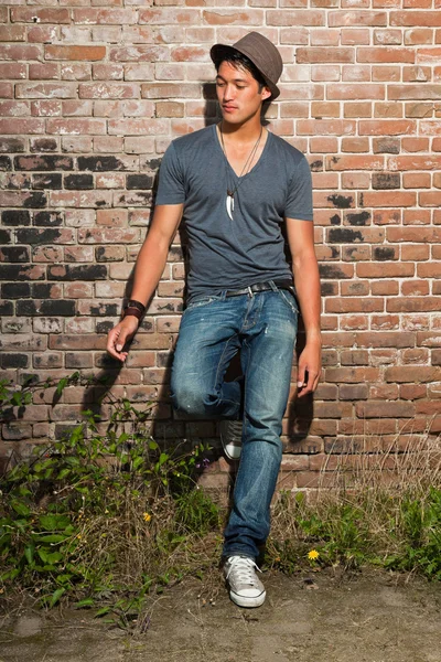 Urban asian man. Good looking. Cool guy. Wearing grey shirt and hat and jeans. Standing in front of brick wall. — Stock Photo, Image