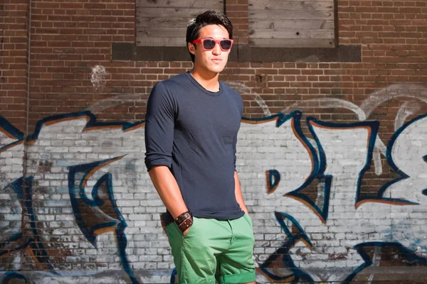 Urban uomo asiatico con occhiali da sole rossi. Bell'aspetto. Che figo. Indossa camicia blu scuro e pantaloncini verdi. In piedi davanti al muro di mattoni con graffiti . — Foto Stock