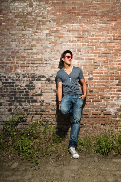 Urbano asiático homem com óculos de sol vermelhos. Boa aparência. Um tipo fixe. Vestindo camisa cinza e chapéu e jeans. Em pé na frente da parede de tijolo . — Fotografia de Stock