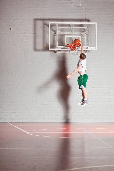 Tvrdý zdravý mladý muž hraje basketbal v tělocvičně krytého. nosí bílé košile a zelené šortky. — Stock fotografie