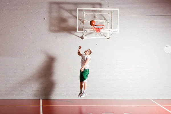 Duro joven saludable jugando baloncesto en el gimnasio interior. Vestir camisa blanca y pantalones cortos verdes . — Foto de Stock