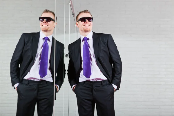 Cool hombre de negocios bien parecido con gafas de sol oscuras de pie contra el espejo . — Foto de Stock