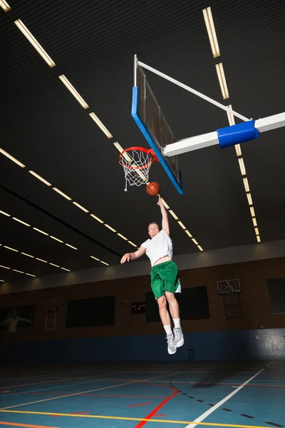 Difficile sano giovane uomo che gioca a basket in palestra al coperto. Indossare camicia bianca e pantaloncini verdi . — Foto Stock