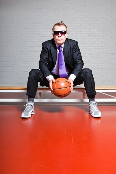 Geschäftsmann mit Basketball. Er trug eine dunkle Sonnenbrille. gut aussehender junger Mann mit kurzen blonden Haaren. Sitzen auf Bank in Turnhalle. — Stockfoto