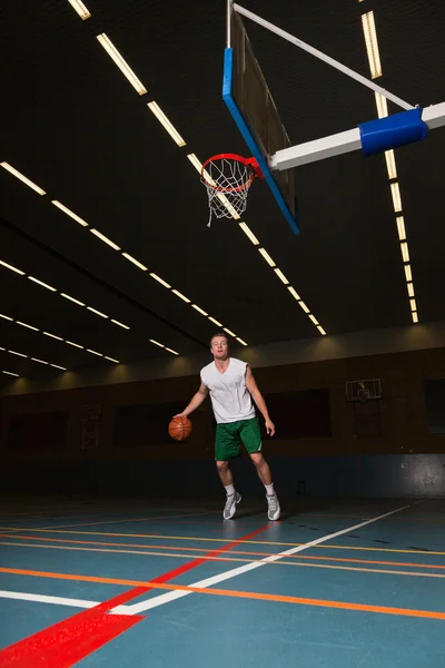 Ein kräftiger, gesunder junger Mann spielt Basketball in der Sporthalle. trägt weißes Hemd und grüne Shorts. — Stockfoto