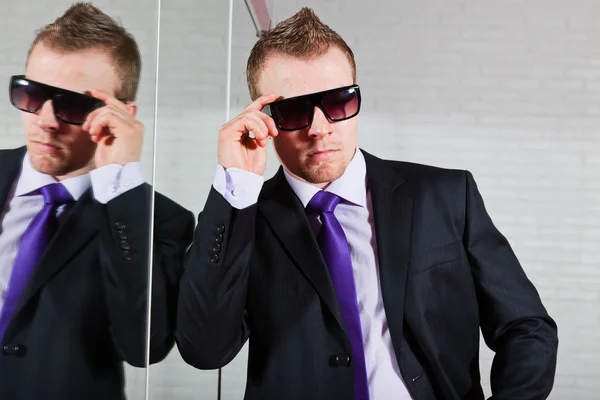Крутой бизнесмен в темных солнцезащитных очках стоит перед зеркалом . — стоковое фото