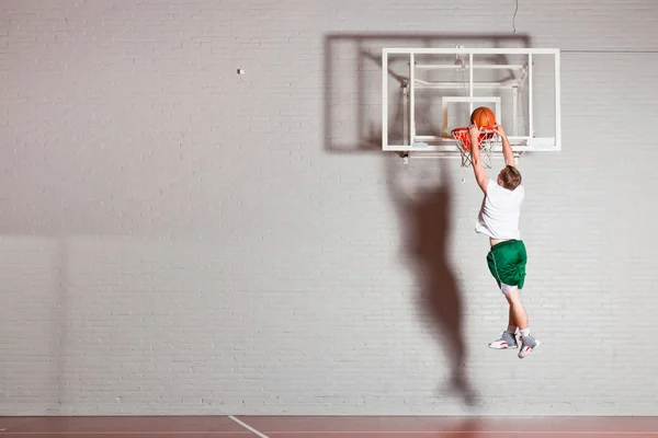 Крутой здоровый молодой человек играет в баскетбол в спортзале. Носит белую рубашку и зеленые шорты . — стоковое фото