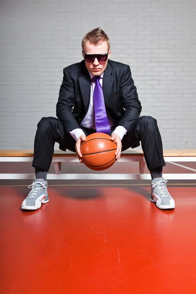 Obchodní muž s basketbal. tmavé brýle. dobře vypadající mladý muž s krátkými blond vlasy. sedí na lavičce v tělocvičně krytého. — Stock fotografie