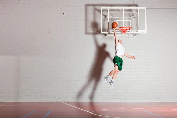 Σκληρή υγιή νεαρό άνδρα που παίζει μπάσκετ στο γυμναστήριο εσωτερική. φορώντας άσπρο πουκάμισο και πράσινο σορτς. — Φωτογραφία Αρχείου