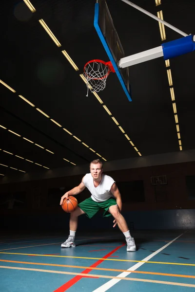 Jovem saudável duro jogando basquete no ginásio indoor. Vestindo camisa branca e shorts verdes . — Fotografia de Stock