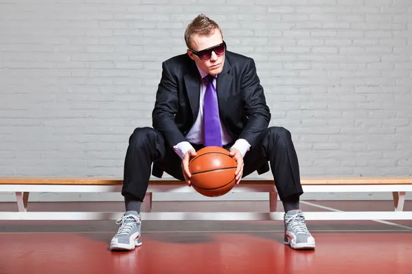 Hombre de negocios con baloncesto. Llevando gafas de sol oscuras. Joven guapo con el pelo corto y rubio. Sentado en el banco en el gimnasio interior . —  Fotos de Stock