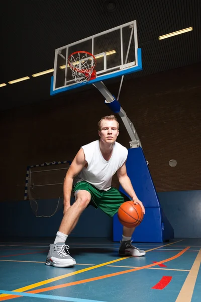 Jovem saudável duro jogando basquete no ginásio indoor. Vestindo camisa branca e shorts verdes . — Fotografia de Stock