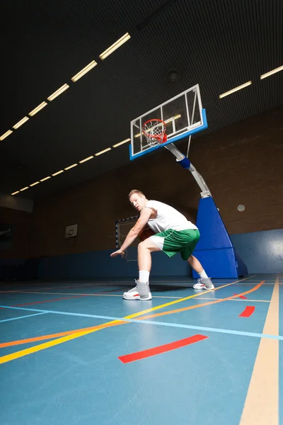 Ein kräftiger, gesunder junger Mann spielt Basketball in der Sporthalle. trägt weißes Hemd und grüne Shorts. — Stockfoto