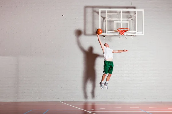 Difficile sano giovane uomo che gioca a basket in palestra al coperto. Indossare camicia bianca e pantaloncini verdi . — Foto Stock