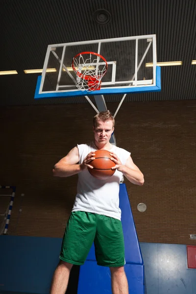 Σκληρή υγιή νεαρό άνδρα που παίζει μπάσκετ στο γυμναστήριο εσωτερική. φορώντας άσπρο πουκάμισο και πράσινο σορτς. — Φωτογραφία Αρχείου