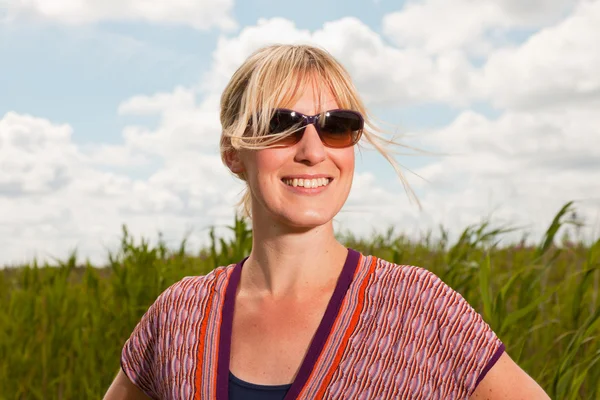 Glad ganska ung blond kvinna med solglasögon njuter av naturen. fält med högt gräs. blå mulen himmel. klädd i rött. — Stockfoto