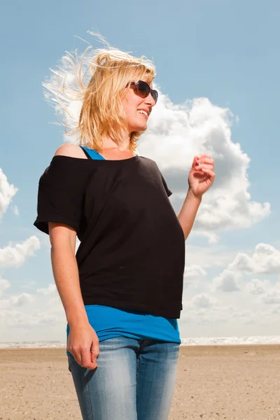 stock image Happy pretty blond woman on the beach. Enjoying nature. Blue cloudy sky. Wearing black sweater and sunglasses.