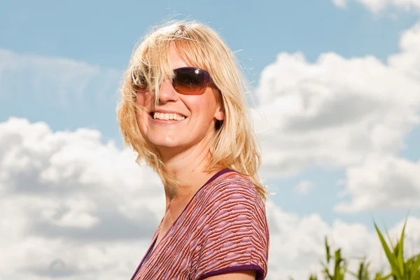 Glückliche junge blonde Frau mit Sonnenbrille, die die Natur genießt. Feld mit hohem Gras. blauer bewölkter Himmel. gekleidet in rot. — Stockfoto