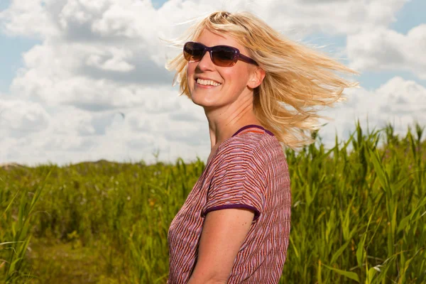 Joyeux jeune femme blonde avec des lunettes de soleil profitant de la nature. Champ avec herbe haute. Ciel nuageux bleu. Habillé en rouge . — Photo