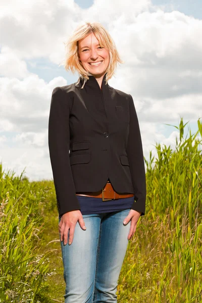 Glückliche junge blonde Frau, die die Natur genießt. Feld mit hohem Gras. blauer bewölkter Himmel. schwarze Jacke. — Stockfoto