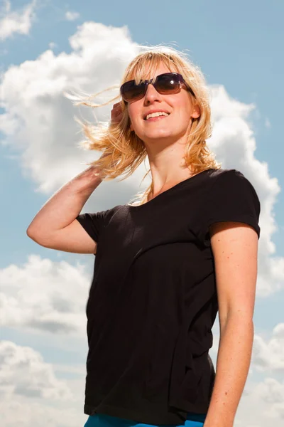 Feliz mujer rubia bonita en la playa. Disfrutando de la naturaleza. Cielo nublado azul. El uso de suéter negro y gafas de sol . — Foto de Stock