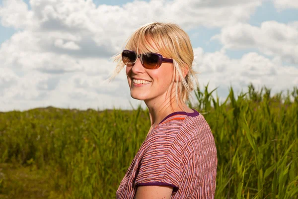 Zadowolony ładny blond kobieta z okulary z przyrodą. pole z wysokiej trawy. błękitne niebo pochmurne. ubrany w czerwony. — Zdjęcie stockowe